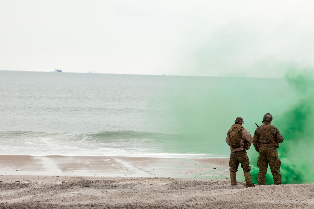 Two military personnel in camouflage uniforms stand on a sandy beach facing the ocean, surrounded by bright green smoke, likely from a smoke grenade or signal device. The scene appears to be a military exercise or training operation, with a vessel visible on the horizon. The green smoke is being used as a visual signal marker.