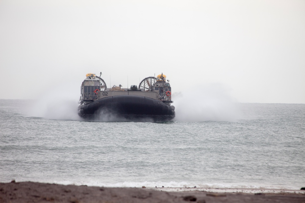 A military hovercraft, marked with the number '02', is approaching a shoreline over open water. The craft is generating significant spray and mist as it travels at speed on its air cushion. The overcast sky and hazy conditions suggest an amphibious landing exercise or operational deployment.