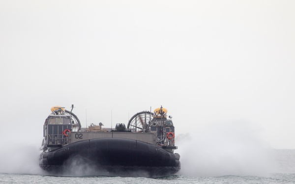 A military hovercraft, marked with the number '02', is approaching a shoreline over open water. The craft is generating significant spray and mist as it travels at speed on its air cushion. The overcast sky and hazy conditions suggest an amphibious landing exercise or operational deployment.