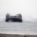 A military hovercraft, marked with the number '02', is approaching a shoreline over open water. The craft is generating significant spray and mist as it travels at speed on its air cushion. The overcast sky and hazy conditions suggest an amphibious landing exercise or operational deployment.