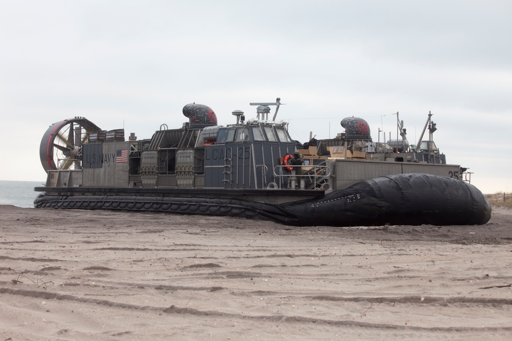A U.S. Navy Landing Craft Air Cushion (LCAC) designated LCAC-25 is beached on a sandy shoreline. The hovercraft is resting on its deflated skirt on the beach, with personnel visible on deck. Military vehicles appear to be loaded on the craft's cargo deck.