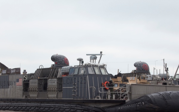 A U.S. Navy Landing Craft Air Cushion (LCAC) designated LCAC-25 is beached on a sandy shoreline. The hovercraft is resting on its deflated skirt on the beach, with personnel visible on deck. Military vehicles appear to be loaded on the craft's cargo deck.