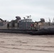 A U.S. Navy Landing Craft Air Cushion (LCAC) designated LCAC-25 is beached on a sandy shoreline. The hovercraft is resting on its deflated skirt on the beach, with personnel visible on deck. Military vehicles appear to be loaded on the craft's cargo deck.