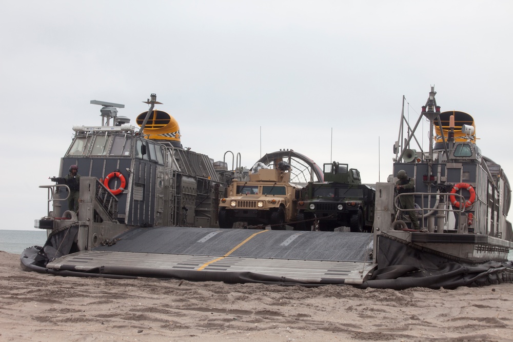 A Landing Craft Air Cushion (LCAC) hovercraft is beached on a sandy shoreline with its bow ramp lowered, carrying military vehicles including at least one tan HMMWV (Humvee) and one olive-drab military truck. Two operator cab structures are visible on either side of the craft, each equipped with orange life rings, yellow radar/antenna equipment, and personnel. The scene appears to depict an amphibious landing operation or exercise.