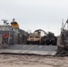 A Landing Craft Air Cushion (LCAC) hovercraft is beached on a sandy shoreline with its bow ramp lowered, carrying military vehicles including at least one tan HMMWV (Humvee) and one olive-drab military truck. Two operator cab structures are visible on either side of the craft, each equipped with orange life rings, yellow radar/antenna equipment, and personnel. The scene appears to depict an amphibious landing operation or exercise.