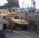 Military personnel are securing a tan/desert-colored HMMWV (Humvee) on what appears to be a naval landing craft or ship deck using chains. At least four service members in different uniforms are visible, working to tie down or release the vehicle. The setting appears to be a military amphibious operation or vehicle loading/offloading exercise.