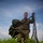 A U.S. Marine in MARPAT digital camouflage uniform is standing in a grassy field, gathering and recovering parachute equipment after a drop. He is holding bundled suspension lines and risers in his right hand while cradling the collapsed parachute canopy under his left arm. The low-angle shot against a partly cloudy blue sky emphasizes the scale and activity of post-jump recovery operations.