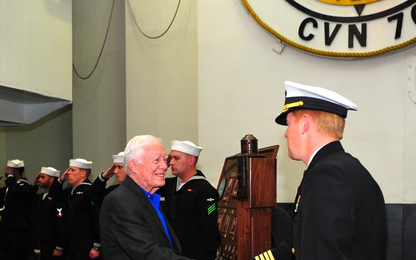 An elderly civilian in a dark blazer and blue shirt is shaking hands with a senior U.S. Navy officer in dress uniform aboard a naval vessel designated CVN 70. A row of enlisted sailors in dress whites stand at attention or salute in the background. The setting appears to be a formal reception or boarding ceremony on the hangar deck of the aircraft carrier.