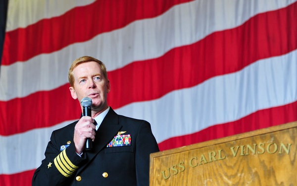 A U.S. Navy officer in dress blues is speaking at a podium aboard the USS Carl Vinson (CVN-70), a Nimitz-class aircraft carrier. The officer holds a handheld microphone and displays multiple service ribbons and aviator wings on his uniform. A large American flag serves as the backdrop for the event.