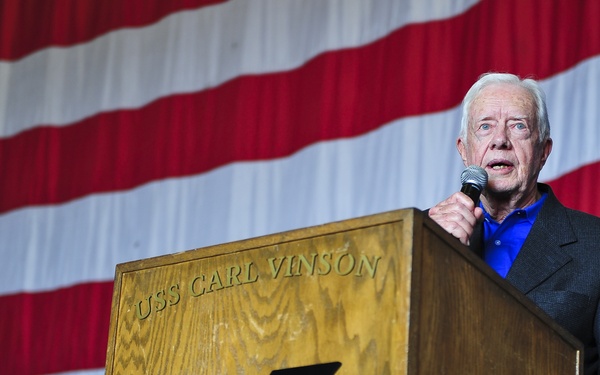An elderly man is speaking at a podium aboard the USS Carl Vinson (CVN-70), a U.S. Navy aircraft carrier. He is holding a microphone and addressing an audience in front of a large American flag backdrop. The podium bears the official emblem of the USS Carl Vinson with a golden eagle and shield insignia.