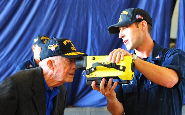 A uniformed military or Coast Guard personnel member is demonstrating a yellow handheld device to an elderly civilian visitor wearing a navy cap. The interaction appears to take place in a hangar or large indoor military facility with blue curtain/tarp backdrop. A second uniformed individual is visible in the background.
