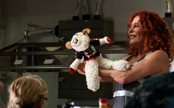 A woman with curly red hair is holding up a stuffed animal toy (appearing to be a lamb or sheep) while smiling, seemingly presenting or entertaining a group of people seated at a table. The seated individuals are wearing what appears to be military camouflage uniforms, suggesting a military dining facility or mess hall setting. The scene has the appearance of a morale-boosting or entertainment visit.