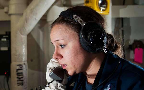 A female U.S. Navy sailor is shown in profile view aboard a naval vessel, speaking into a sound-powered telephone handset while wearing communication headphones. She is dressed in a Navy working uniform with white protective gloves, suggesting she is performing a duty station role, likely during a drill or operational watch. The setting appears to be an interior compartment of a warship.