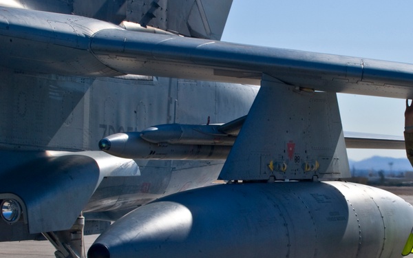 A military aircraft ground crew member in camouflage uniform and high-visibility safety vest is conducting a pre-flight or post-flight inspection of a military jet aircraft on a tarmac. The technician is reaching up to inspect the underside of the wing, wearing hearing protection headset and sunglasses. The aircraft has an external fuel tank (drop tank) and landing gear visible in the foreground.