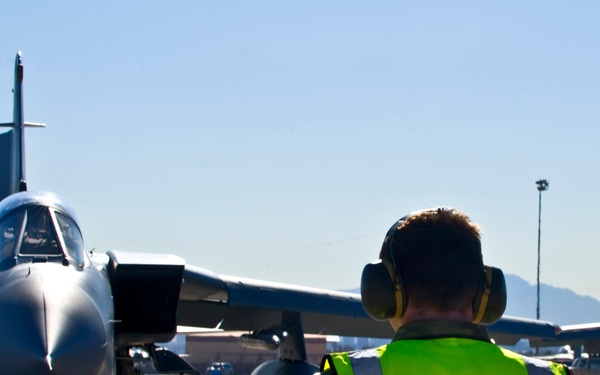 A ground crew member wearing a high-visibility yellow vest and hearing protection is marshaling a military jet aircraft on a flight line apron. The aircraft, which appears to be an F-15 or similar twin-engine tactical jet, is taxiing toward the marshal. The scene takes place at a military airbase with mountains visible in the background.