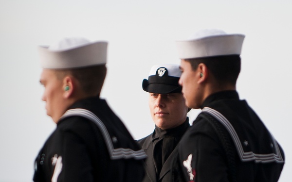 Three U.S. Navy personnel in dress uniform are standing at attention or in formation, likely during a formal ceremony. Two sailors in the foreground wear traditional white 'Dixie cup' sailor caps and dark dress uniforms with neckerchiefs, while the central figure wears a dark officer or petty officer combination cap with a USN eagle insignia. The background suggests an outdoor maritime setting, possibly aboard a ship or near open water.
