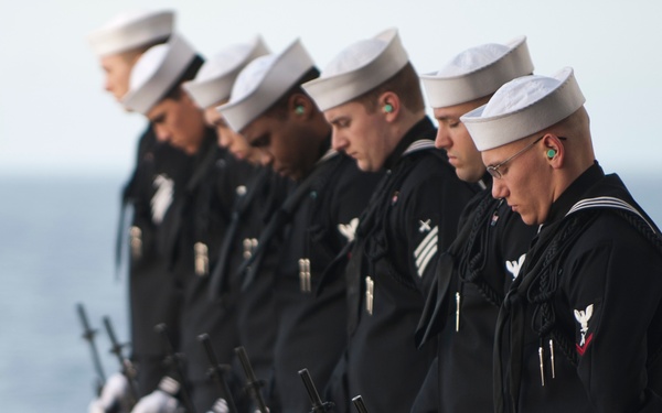 A rifle party of U.S. Navy sailors in dress blue uniforms stands in a solemn line holding rifles pointed downward during what appears to be a burial-at-sea or memorial ceremony. The sailors are positioned on what appears to be the deck of a naval vessel with open ocean visible in the background. Several sailors are wearing ear protection, indicating the rifles will be fired as part of a ceremonial volley.