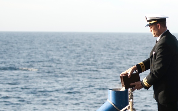 A naval officer in dress uniform stands on the deck of a military vessel, holding what appears to be a wooden urn or box over a blue cylindrical tube near the ship's railing. The scene is consistent with a burial at sea ceremony, with the officer appearing to pour or release cremated remains into the sea. The ocean is visible in the background under an overcast sky.