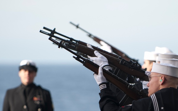 A military burial-at-sea or honors ceremony is taking place aboard a naval vessel over open ocean. A rifle party of U.S. Navy sailors in dress blues and white Dixie cup covers are firing a rifle volley salute. A senior officer or official in a dark dress uniform with medals stands at attention in the background.