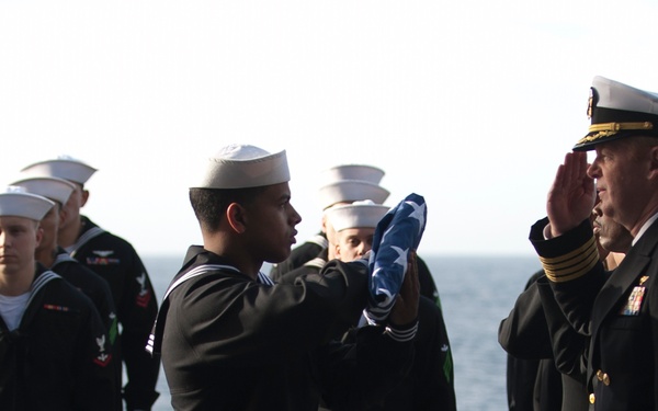A formal U.S. Navy ceremony is taking place on the deck of a naval vessel at sea. A junior enlisted sailor is presenting a folded American flag to a senior officer, who is rendering a salute. Several other sailors in dress uniforms stand at attention in the background.