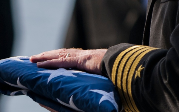 A uniformed military officer is holding a ceremonially folded American flag, consistent with a military funeral or memorial service. The officer's sleeve displays four gold stripes and a gold star, indicating a high-ranking naval or senior military officer. The flag is folded in the traditional triangular shape with the blue field and white stars visible.