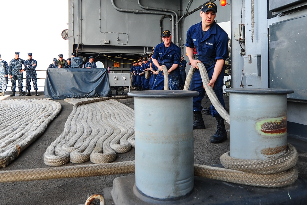 U.S. Navy sailors are engaged in line-handling operations on the deck of a naval vessel, likely during a mooring or departure evolution. A foreground sailor in dark blue Navy working uniform is managing a thick mooring line around a double bollard, while a line of sailors in the background handles the same line in a chain formation. Additional personnel in Navy camouflage uniforms observe from the port side of the deck.