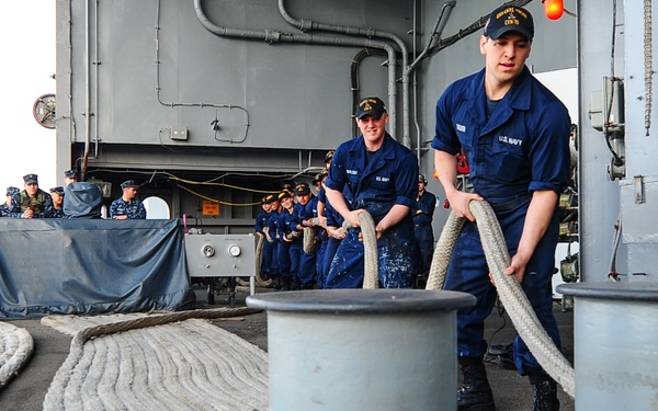 U.S. Navy sailors are engaged in line-handling operations on the deck of a naval vessel, likely during a mooring or departure evolution. A foreground sailor in dark blue Navy working uniform is managing a thick mooring line around a double bollard, while a line of sailors in the background handles the same line in a chain formation. Additional personnel in Navy camouflage uniforms observe from the port side of the deck.