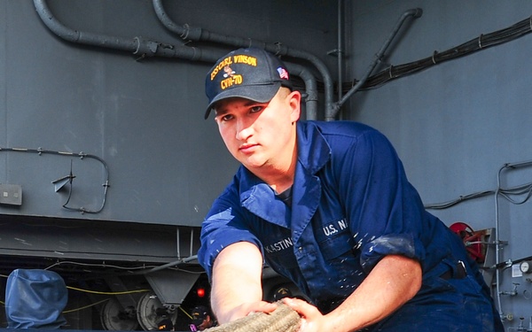 A U.S. Navy sailor assigned to USS Carl Vinson (CVN-70) is pulling a large mooring line on the ship's deck. The sailor is wearing blue Navy coveralls, black combat boots, and a ship's ball cap with 'Carl Vinson CVN-70' visible. Water droplets are spraying from the rope, indicating significant tension and physical exertion during what appears to be a mooring or line-handling evolution.