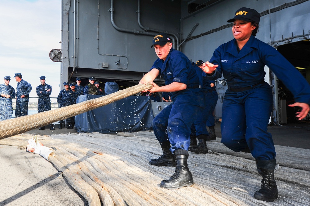 U.S. Navy sailors are hauling a large mooring line on what appears to be the deck of a naval vessel, likely during a port arrival or departure operation. Two sailors in the foreground — one male and one female (name tag reads 'SMITH') — are actively pulling the heavy rope while a group of uniformed personnel observe in the background. The setting is a ship's deck or pier area with naval vessel superstructure visible.