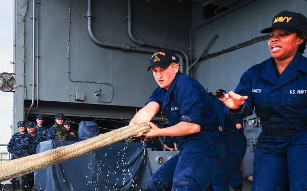 U.S. Navy sailors are hauling a large mooring line on what appears to be the deck of a naval vessel, likely during a port arrival or departure operation. Two sailors in the foreground — one male and one female (name tag reads 'SMITH') — are actively pulling the heavy rope while a group of uniformed personnel observe in the background. The setting is a ship's deck or pier area with naval vessel superstructure visible.