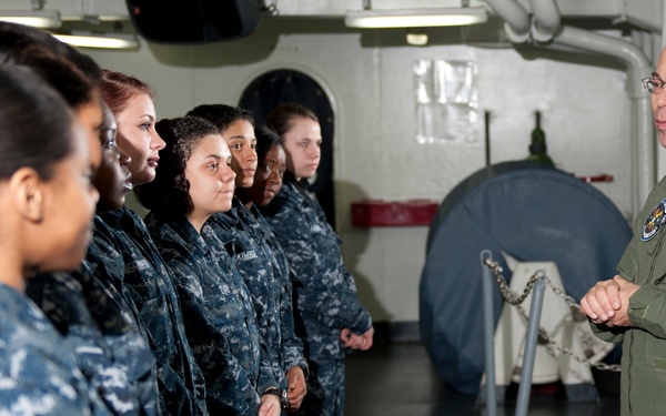 A senior military officer in an olive green flight suit is addressing a group of Navy enlisted personnel wearing Navy Working Uniform (NWU) digital camouflage. The setting appears to be the interior of a naval vessel, likely a briefing or inspection scenario. The officer appears to be speaking to a predominantly female group of sailors standing in formation.