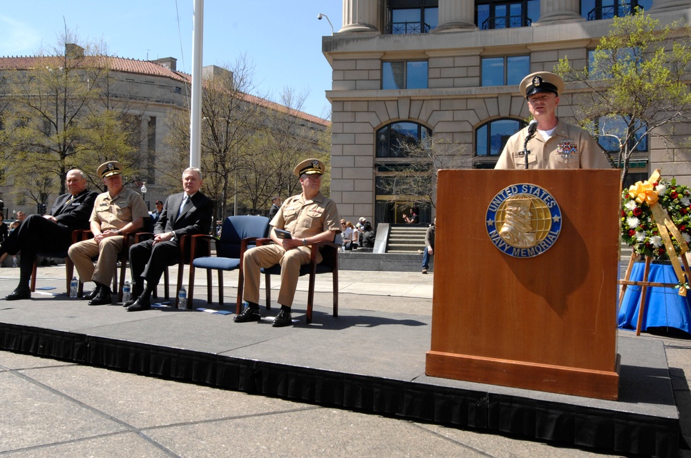 A formal ceremony is taking place at the United States Navy Memorial, likely in Washington D.C. A Navy enlisted sailor in khaki uniform is speaking at the podium while several military officers and a civilian are seated on a raised stage behind him. A floral wreath is visible to the right of the podium.