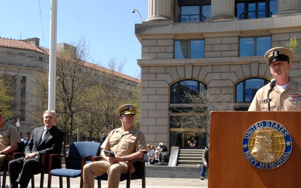 A formal ceremony is taking place at the United States Navy Memorial, likely in Washington D.C. A Navy enlisted sailor in khaki uniform is speaking at the podium while several military officers and a civilian are seated on a raised stage behind him. A floral wreath is visible to the right of the podium.