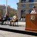 A formal ceremony is taking place at the United States Navy Memorial, likely in Washington D.C. A Navy enlisted sailor in khaki uniform is speaking at the podium while several military officers and a civilian are seated on a raised stage behind him. A floral wreath is visible to the right of the podium.
