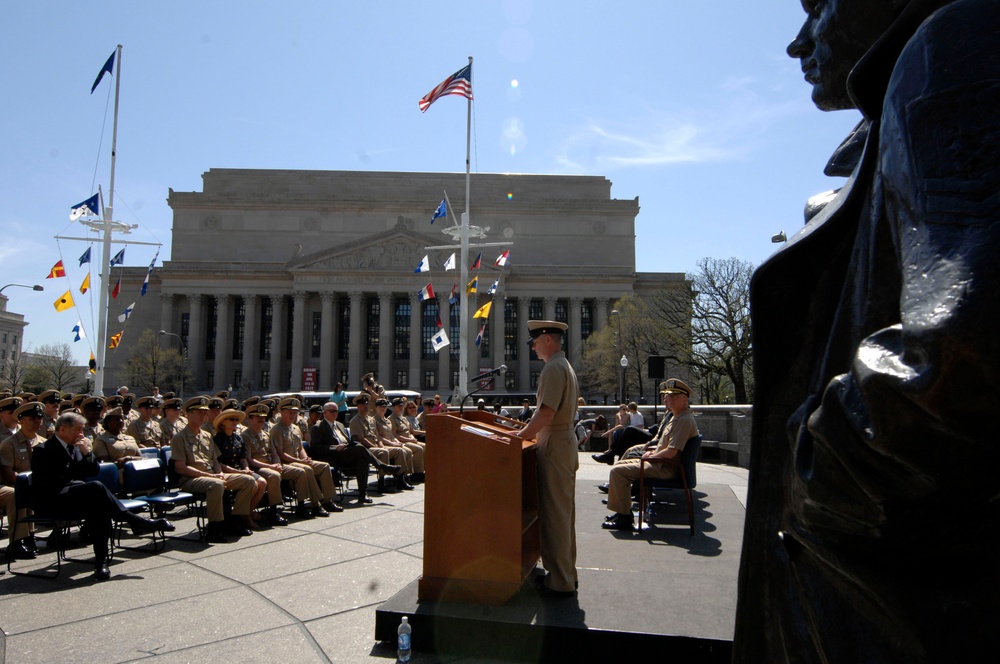 A military ceremony is taking place outdoors in front of a large neoclassical government building, likely the Navy Memorial or a similar Washington D.C. landmark. A uniformed officer stands at a wooden podium addressing rows of seated military personnel in tan/khaki uniforms. Bronze statues are visible in the foreground right, and multiple naval signal flags are displayed on flagpoles along with the American flag.
