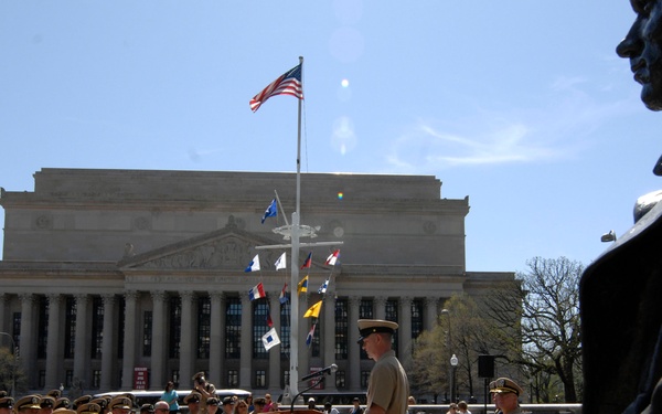 A military ceremony is taking place outdoors in front of a large neoclassical government building, likely the Navy Memorial or a similar Washington D.C. landmark. A uniformed officer stands at a wooden podium addressing rows of seated military personnel in tan/khaki uniforms. Bronze statues are visible in the foreground right, and multiple naval signal flags are displayed on flagpoles along with the American flag.