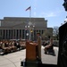 A military ceremony is taking place outdoors in front of a large neoclassical government building, likely the Navy Memorial or a similar Washington D.C. landmark. A uniformed officer stands at a wooden podium addressing rows of seated military personnel in tan/khaki uniforms. Bronze statues are visible in the foreground right, and multiple naval signal flags are displayed on flagpoles along with the American flag.