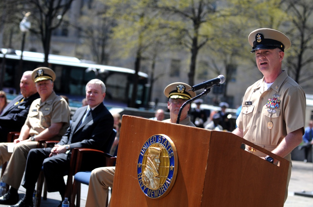 A U.S. Navy enlisted senior chief or master chief petty officer is speaking at a podium bearing the United States Navy Memorial seal at an outdoor ceremony. Several dignitaries and uniformed naval officers are seated in the background. The setting appears to be the U.S. Navy Memorial plaza in Washington, D.C., during spring based on the budding trees.