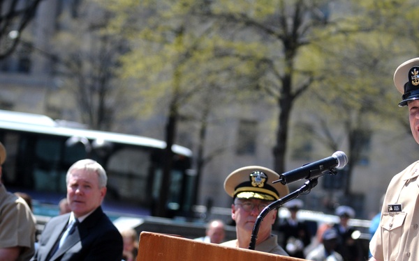 A U.S. Navy enlisted senior chief or master chief petty officer is speaking at a podium bearing the United States Navy Memorial seal at an outdoor ceremony. Several dignitaries and uniformed naval officers are seated in the background. The setting appears to be the U.S. Navy Memorial plaza in Washington, D.C., during spring based on the budding trees.