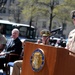 A U.S. Navy enlisted senior chief or master chief petty officer is speaking at a podium bearing the United States Navy Memorial seal at an outdoor ceremony. Several dignitaries and uniformed naval officers are seated in the background. The setting appears to be the U.S. Navy Memorial plaza in Washington, D.C., during spring based on the budding trees.