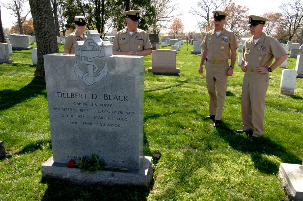 Four U.S. Navy personnel in service khaki uniforms are gathered at the grave of Delbert D. Black at what appears to be Arlington National Cemetery. The headstone identifies him as GMCM U.S. Navy, the First Master Chief Petty Officer of the Navy, a Pearl Harbor Survivor who lived from July 11, 1922 to March 5, 2000. A red rose has been placed at the base of the headstone as a tribute.