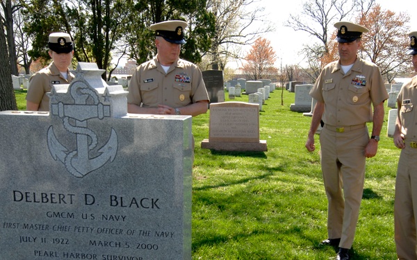 Four U.S. Navy personnel in service khaki uniforms are gathered at the grave of Delbert D. Black at what appears to be Arlington National Cemetery. The headstone identifies him as GMCM U.S. Navy, the First Master Chief Petty Officer of the Navy, a Pearl Harbor Survivor who lived from July 11, 1922 to March 5, 2000. A red rose has been placed at the base of the headstone as a tribute.