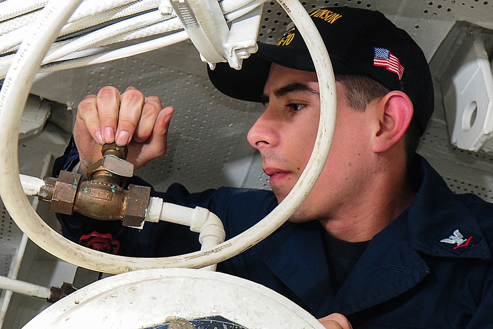 A U.S. Navy sailor is performing maintenance work on piping or valve systems aboard a naval vessel. The sailor is wearing a dark navy uniform with a black cover (cap) displaying an American flag patch and what appears to be a ship name. He is focused on manipulating a brass valve or fitting connected to white PVC-style piping.