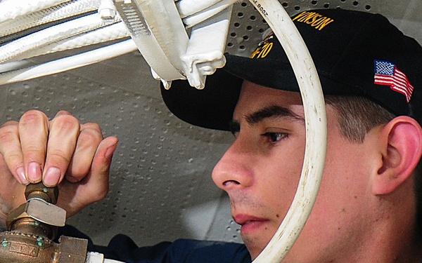 A U.S. Navy sailor is performing maintenance work on piping or valve systems aboard a naval vessel. The sailor is wearing a dark navy uniform with a black cover (cap) displaying an American flag patch and what appears to be a ship name. He is focused on manipulating a brass valve or fitting connected to white PVC-style piping.
