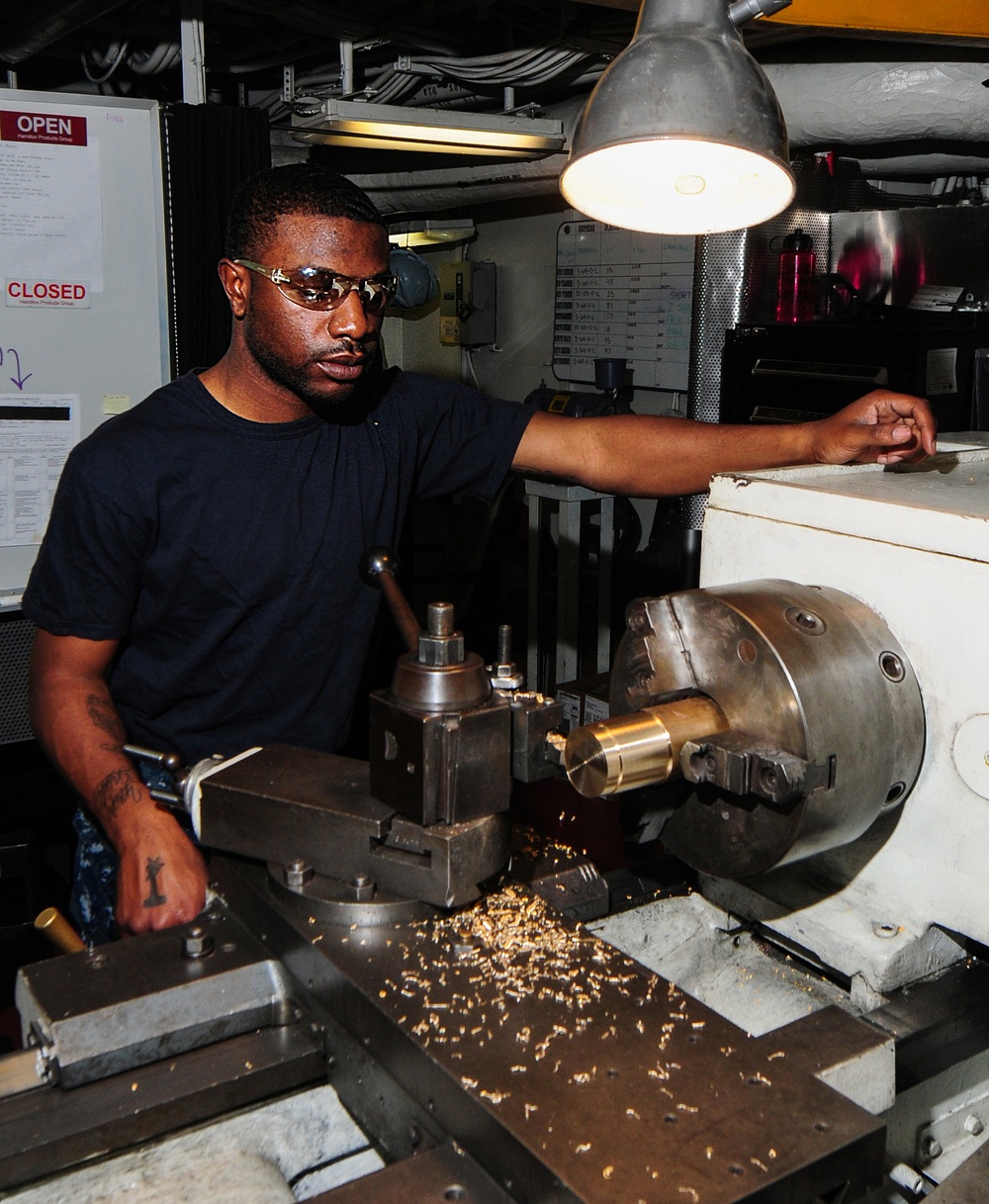 A man wearing safety glasses and a dark navy t-shirt operates a metal lathe in what appears to be a machine shop or workshop environment. Brass or gold-colored metal shavings are scattered across the lathe bed, indicating active machining of a brass workpiece secured in the chuck. The setting, with its overhead piping, cable runs, and posted signs, suggests an industrial or military vessel machine shop.