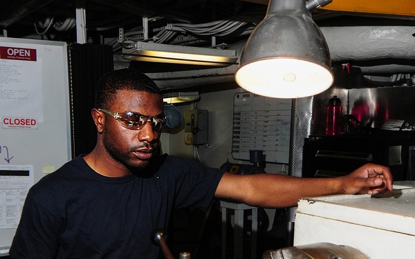 A man wearing safety glasses and a dark navy t-shirt operates a metal lathe in what appears to be a machine shop or workshop environment. Brass or gold-colored metal shavings are scattered across the lathe bed, indicating active machining of a brass workpiece secured in the chuck. The setting, with its overhead piping, cable runs, and posted signs, suggests an industrial or military vessel machine shop.