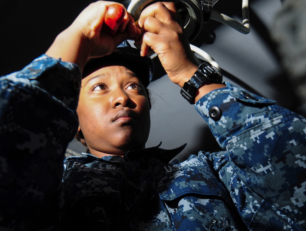 A U.S. Navy sailor in Navy Working Uniform (NWU) digital camouflage is performing maintenance or repair work on overhead equipment. The sailor is looking upward while using a red-handled tool, working on what appears to be a mechanical component. The low-angle shot provides a dramatic perspective of the work being performed.