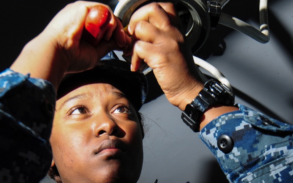 A U.S. Navy sailor in Navy Working Uniform (NWU) digital camouflage is performing maintenance or repair work on overhead equipment. The sailor is looking upward while using a red-handled tool, working on what appears to be a mechanical component. The low-angle shot provides a dramatic perspective of the work being performed.