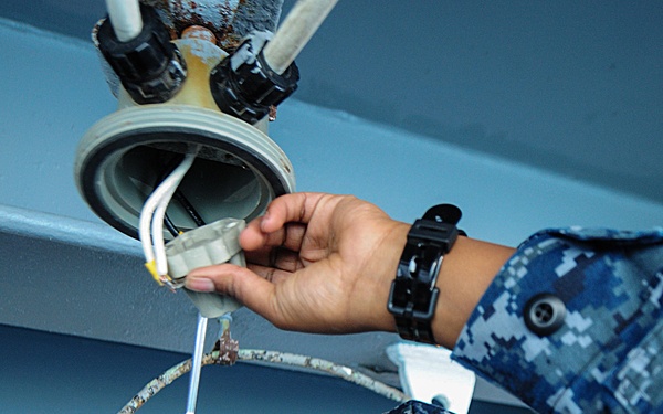 A U.S. Navy sailor in Navy Working Uniform (NWU) Type I digital camouflage is performing electrical maintenance work overhead, using a red-handled screwdriver on a ceiling-mounted light fixture or electrical component. The sailor is looking upward with focused concentration while working on exposed wiring. A second pair of hands, also in NWU uniform, is visible assisting with the task from above.