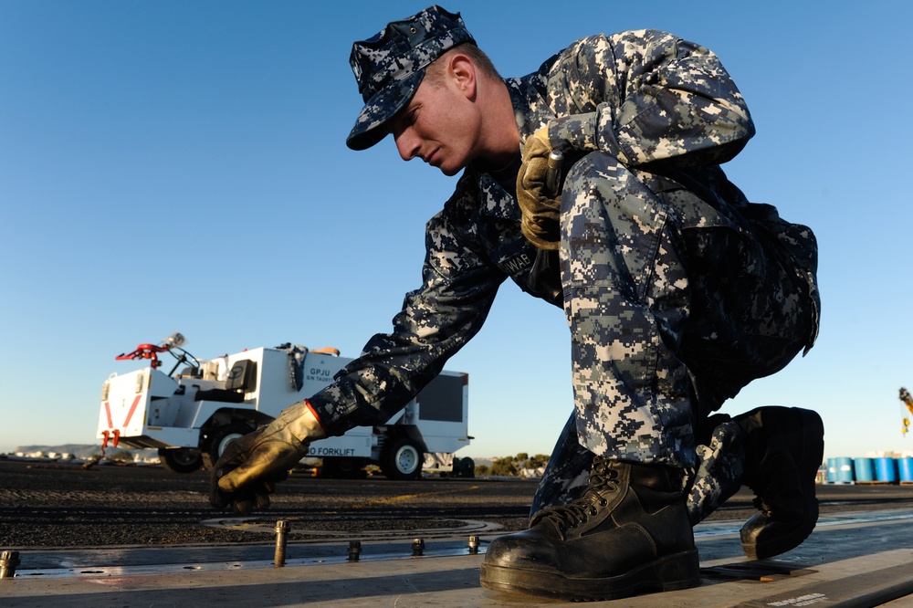 A U.S. Navy sailor in Navy Working Uniform (NWU) Type I digital camouflage is crouching down on what appears to be a flight deck or tarmac, performing a hands-on maintenance or inspection task. The sailor is wearing gold-colored work gloves and black combat boots. A white ground support vehicle is visible in the background on the airfield.